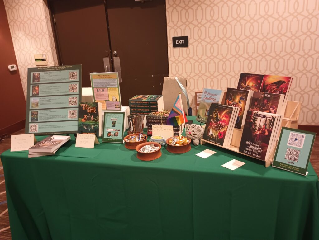 A photo of a book sale table. A large sign anchors the left side, and a stairstep-style display anchors the right side. Between these are various signs, stacks of books, bowls of candy and pins, and a small plushie cat holding a Pride flag.